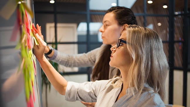 Zwei Frauen stehen vor einem Flipchart und kleben Post-its darauf