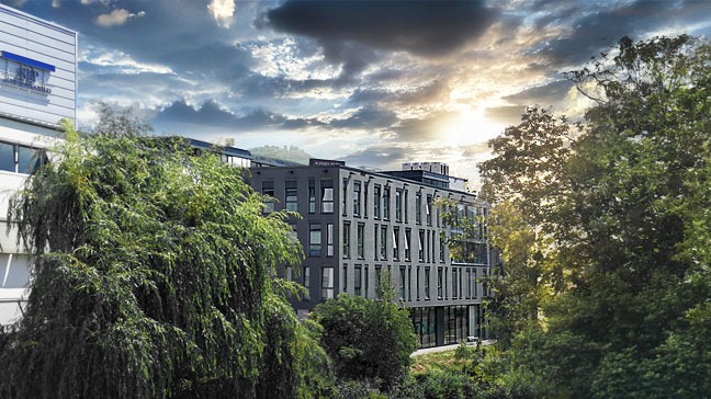 Modernes Firmengebäude umgeben von Bäumen und Grünflächen unter einem bewölkten Himmel